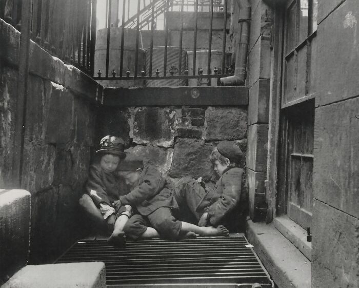 Three children in worn clothing sitting in a dark alley, illustrating 1800s America poverty and hardship.