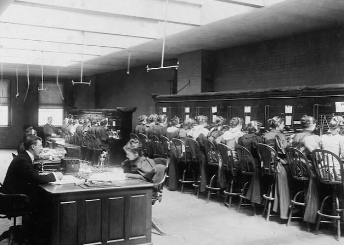 Women working at a telephone switchboard in a large room, showcasing 1800s America communication and labor history.
