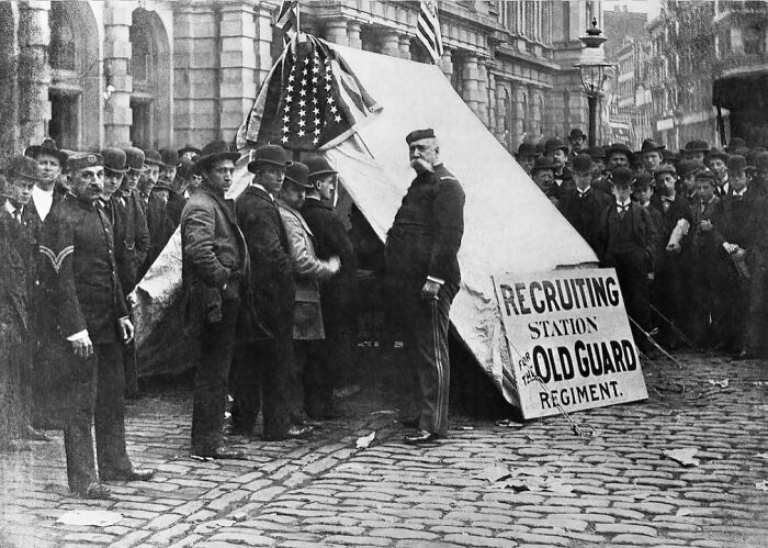 Men in 1800s America gathered around a recruiting station tent for the Old Guard regiment on a cobblestone street.