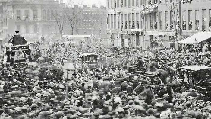 Crowded street scene in 1800s America with horse-drawn carriages and people gathered along city buildings.
