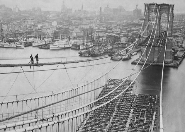 Suspended workers on Brooklyn Bridge cables with 1800s America cityscape and waterfront steamboats in the background.