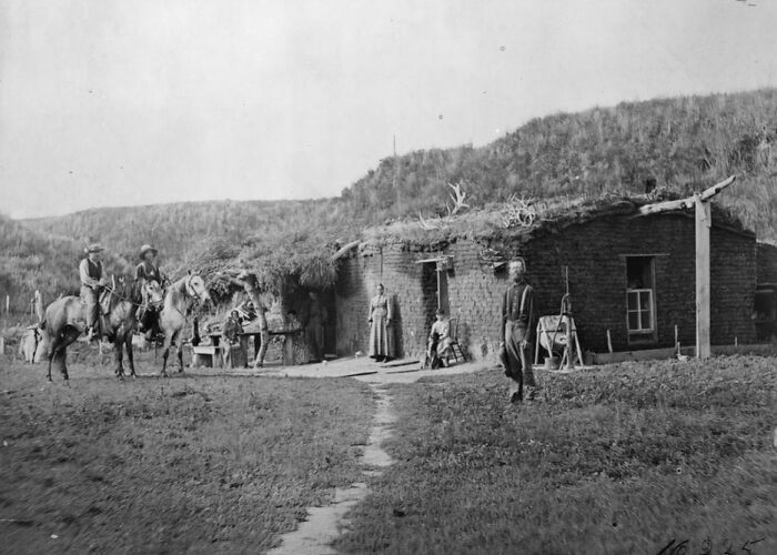 Black and white image of 1800s America showing people, horses, and adobe buildings with grass-covered roofs.