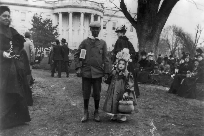Black and white photo of children dressed in 1800s America attire near a historic building with people gathered in the background