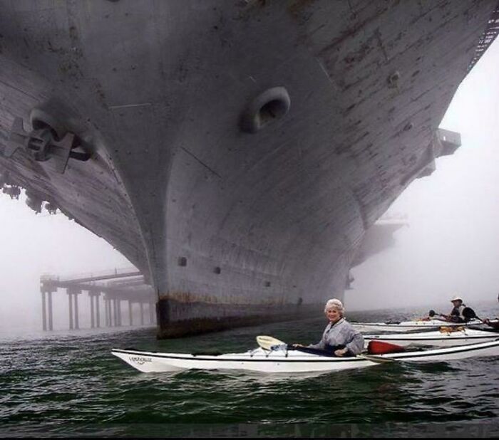 A small kayak with a person paddling near the enormous bow of an extremely large ship emerging from heavy fog, an example of how large things can be eerie.