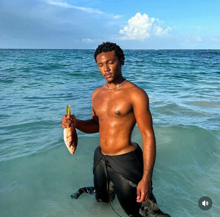 Young man holding a fish standing in the ocean water, illustrating Instagram vs reality moments behind the glam lifestyle.