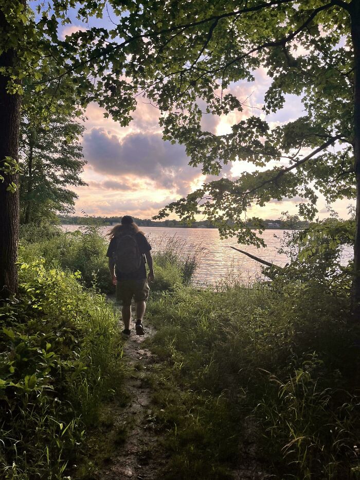 Person walking on a forest path toward a lake at sunset creating beautiful accidental Renaissance photo effect.