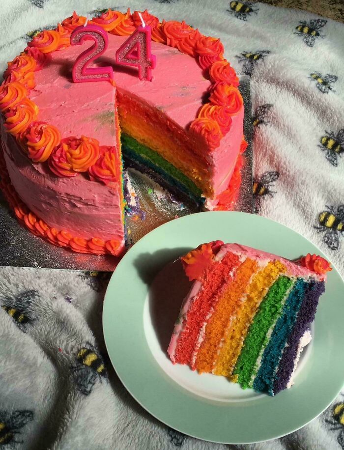 Rainbow layered baked goods birthday cake with pink and orange frosting, slice served on a plate on a bee-patterned blanket.