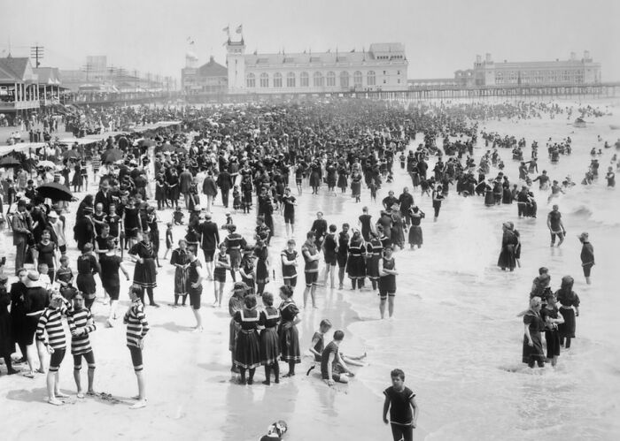 Crowded 1800s America beach scene with people in period bathing suits enjoying the water and sand on a sunny day.