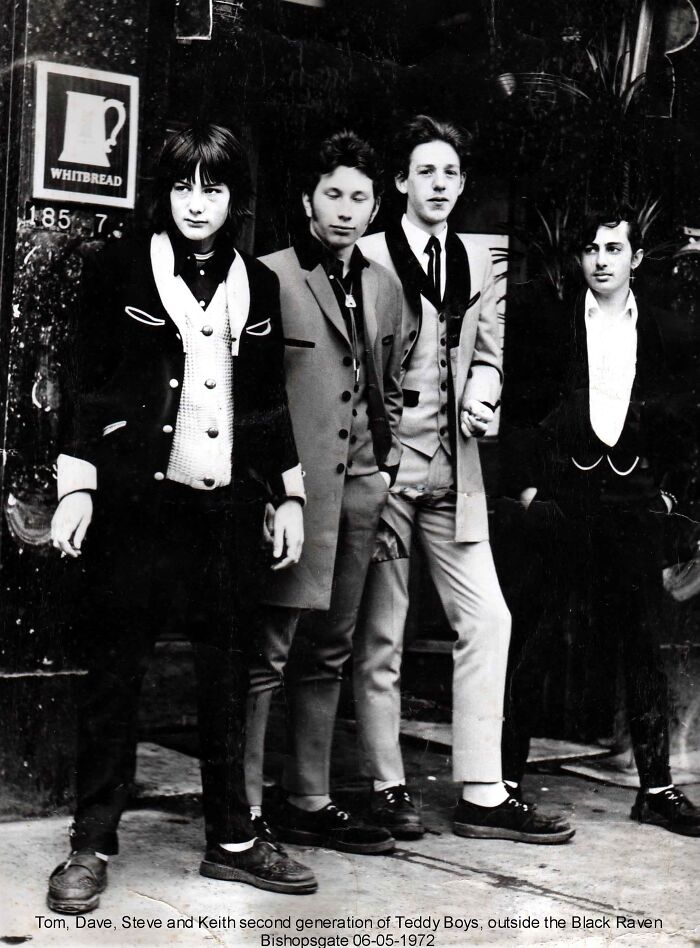 Four young men dressed in vintage suits representing the 20th century Teddy Boy era standing outside a pub.