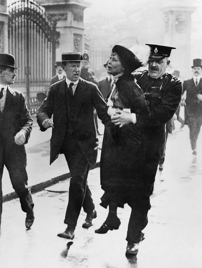 Woman being arrested by police during 20th century suffrage era protest, surrounded by men in formal attire on street.