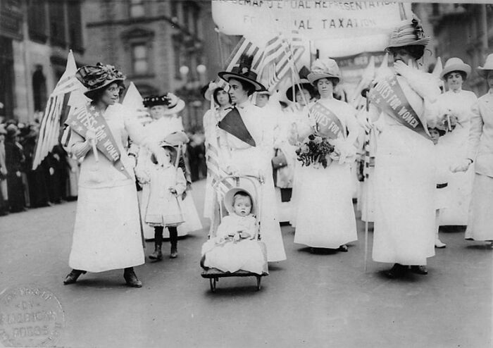 Women in the 20th century suffrage era march in white dresses and sashes, holding flags and banners advocating voting rights.