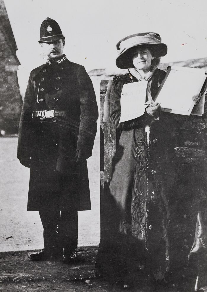 Woman in early 20th century dress holding suffrage petition, standing next to a police officer during suffrage era protest.