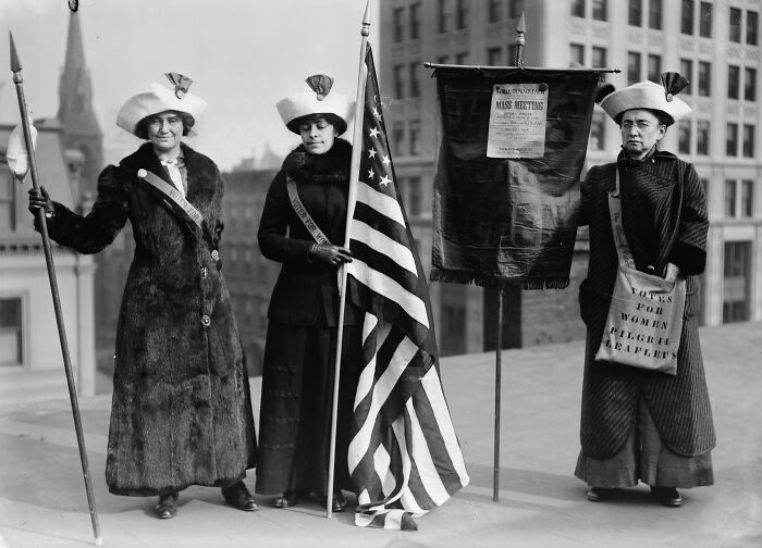 Three women in suffrage-era clothing holding flags and banners during a powerful 20th century women suffrage protest.