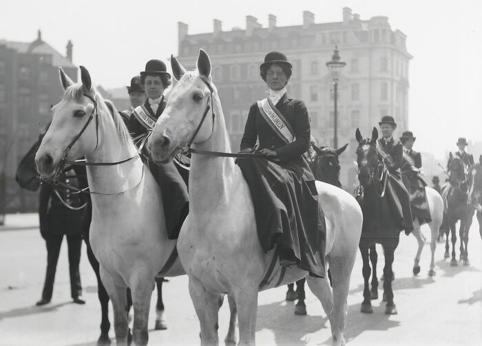 Women in the 20th century suffrage era wearing sashes riding horses during a historic protest march.