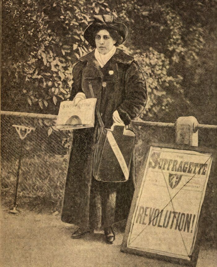 Woman in 20th century suffrage era attire, holding papers next to a suffragette revolution sign in a historic black and white photo.