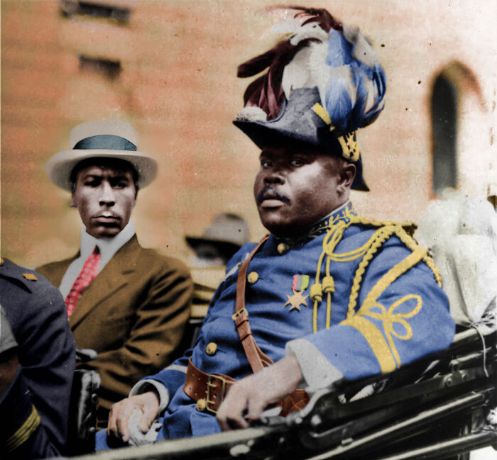 African American man in decorated military uniform and feathered hat seated beside man in suit and white hat, 1920s colorized photo.