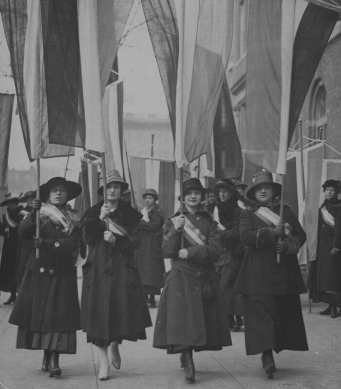Women marching with flags during the 20th century suffrage era wearing coats and hats in a historic protest scene.