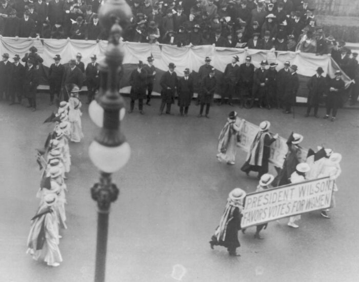 Women marching in a suffrage era protest holding a banner supporting votes for women near a crowd of onlookers in the 20th century
