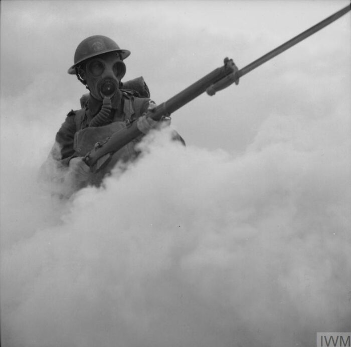 Black and white historical photo of a soldier in a gas mask holding a rifle emerging through thick smoke.