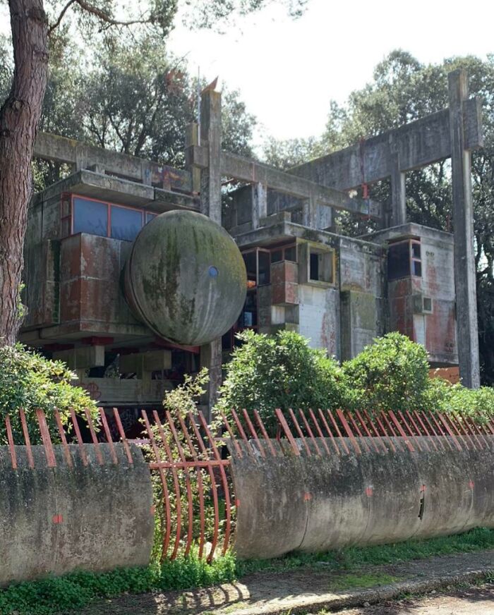 Abandoned concrete structure with rusted metal bars and overgrown greenery in a forested area, showing decay and neglect.