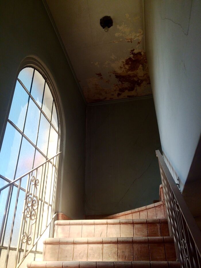 Abandoned places interior showing worn staircase, c*****d walls, rusty ceiling, and a large arched window with natural light.
