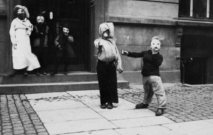 Children wearing unusual masks and costumes on a cobblestone street in a weird and wild historical photo.