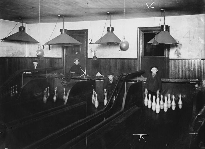 Black and white historical photo of boys playing early bowling in a vintage wooden bowling alley with numbered lanes.