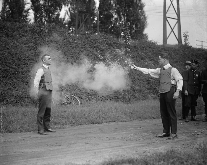 Two men in bulletproof vests testing gunfire outdoors in a weird historical photo with police watching nearby.