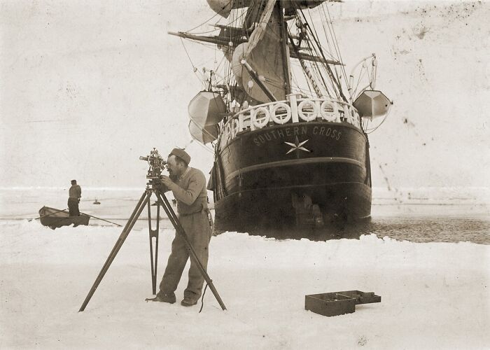 Historical photo of a man using surveying equipment on ice near the ship Southern Cross in a frozen landscape.