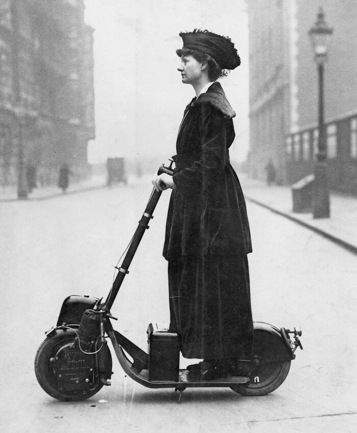 Woman in early 20th century clothing riding a vintage motorized scooter in a historical photo.