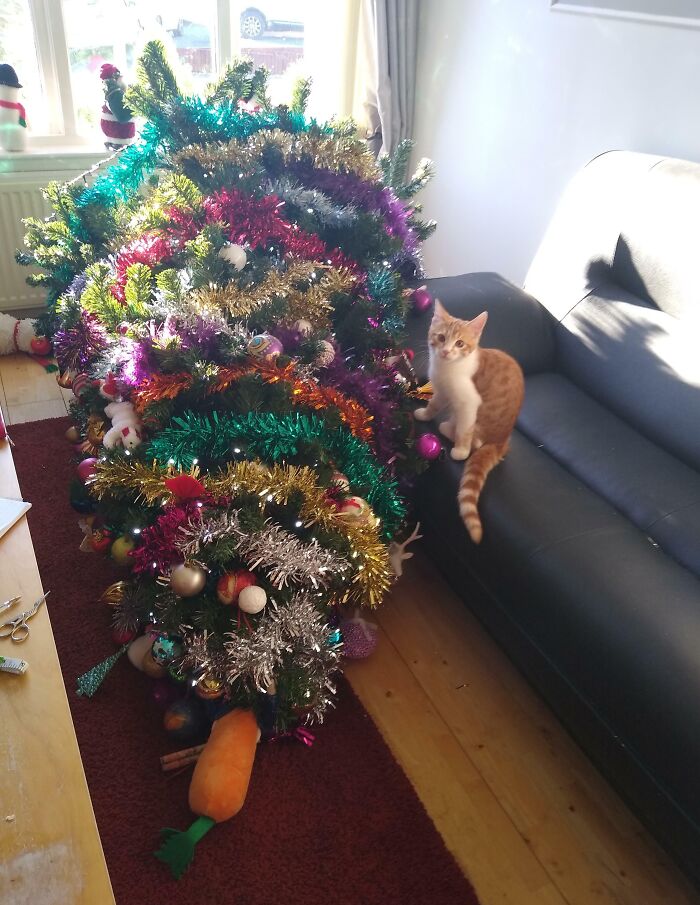 Cat causing chaos by knocking over a decorated Christmas tree covered in colorful tinsel and ornaments.