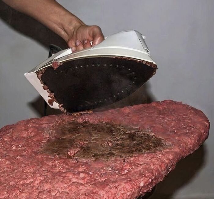 Hand holding an iron with burnt residue pressing down on a large, raw ground meat surface unhealthy food concept.