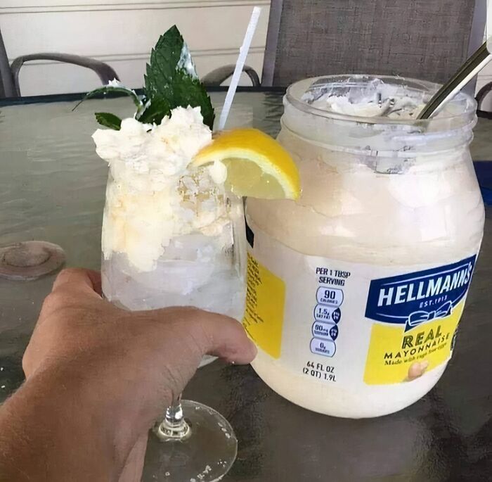 Hand holding a cocktail glass with mayonnaise garnish next to a large jar of Hellmann's real mayonnaise, unhealthy food concept.
