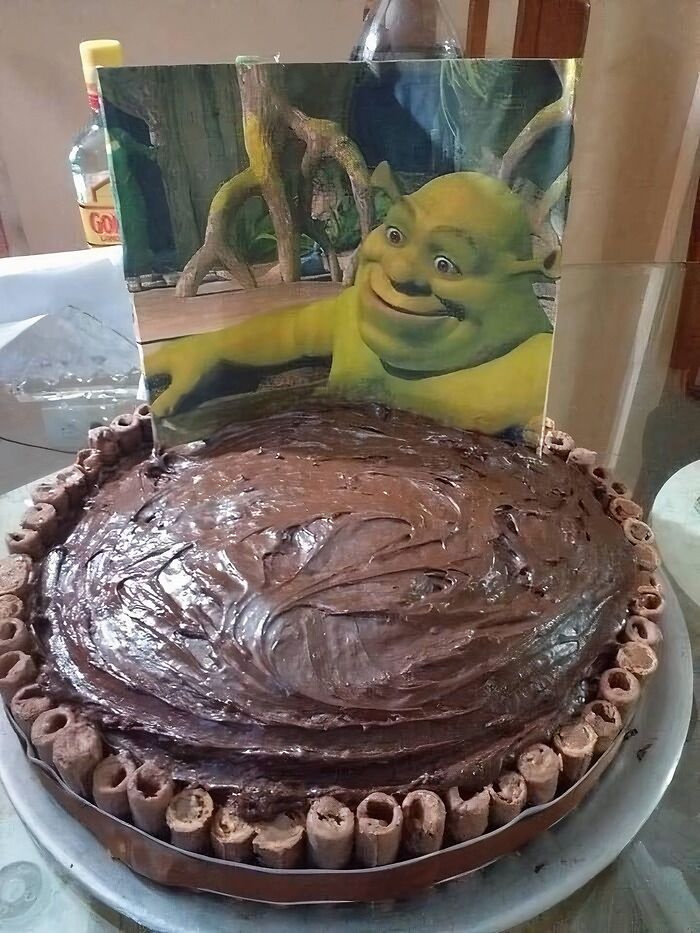 Chocolate cake topped with chocolate sticks on a plate, featuring a Shrek character cutout in the background.