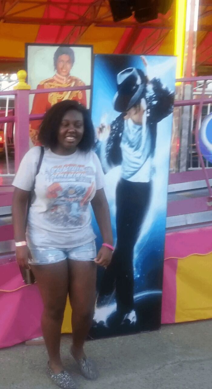 Young woman smiling next to a Michael Jackson poster at a carnival, capturing a fun moment from her regrettable past.
