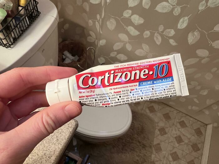Hand holding a tube of hydrocortisone cream in a bathroom, illustrating funny ways people learned their lesson.