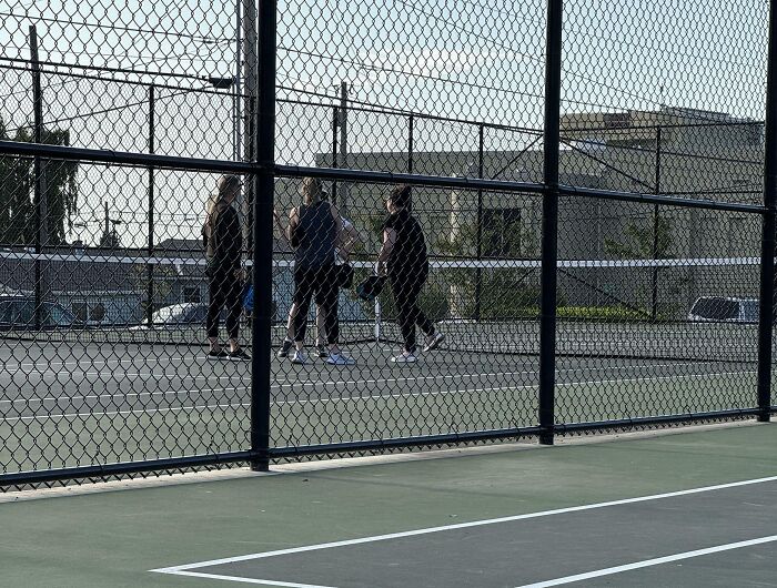 People walking and standing on a tennis court behind a fence, ignoring rules and showing no care about etiquette.