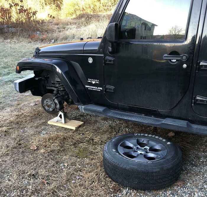 Black Jeep Wrangler on jack stand with missing front tire and tire lying on ground, illustrating funny ways people learned their lesson.