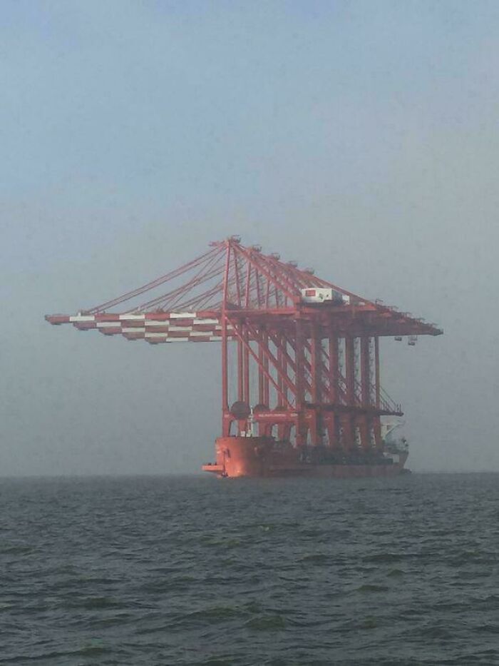 An extremely large red gantry crane being transported on a ship, almost disappearing into the misty horizon, showing how extremely large things can be eerie.