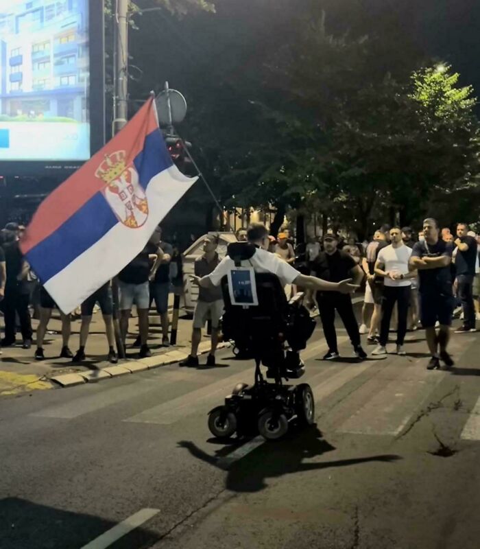 Man in motorized wheelchair waving Serbian flag in front of a crowd, an accidental Renaissance photo capturing a museum-worthy moment.
