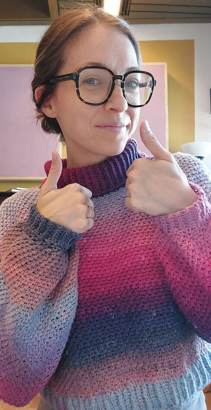 Woman wearing colorful crocheted sweater and black glasses giving thumbs up, showcasing crocheting project skills.