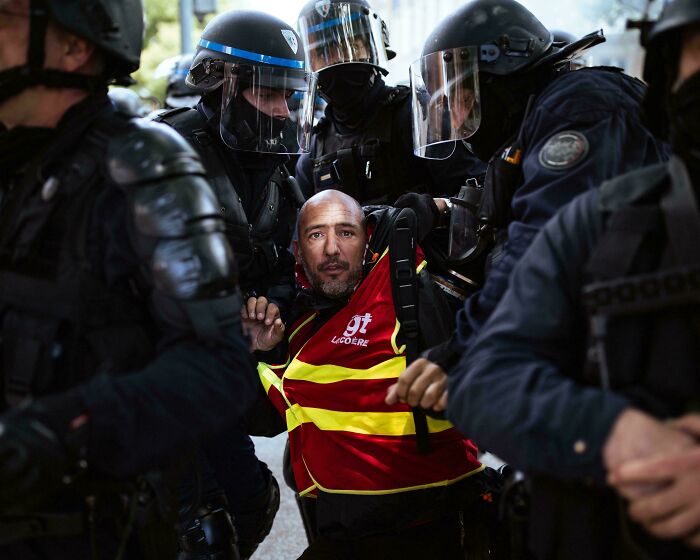 Man in red and yellow vest surrounded by police officers in riot gear, capturing an accidental Renaissance moment in photography.