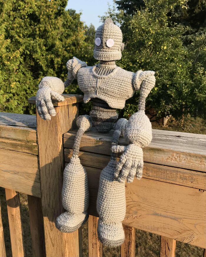 Crocheted robot figure sitting on a wooden fence, showcasing detailed craftsmanship by crocheting enthusiasts outdoors.