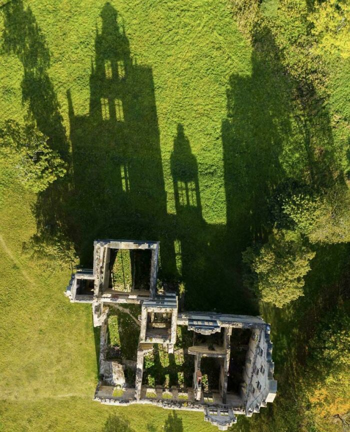 Aerial view of ancient ruins casting shadows resembling a Renaissance masterpiece on green grass.