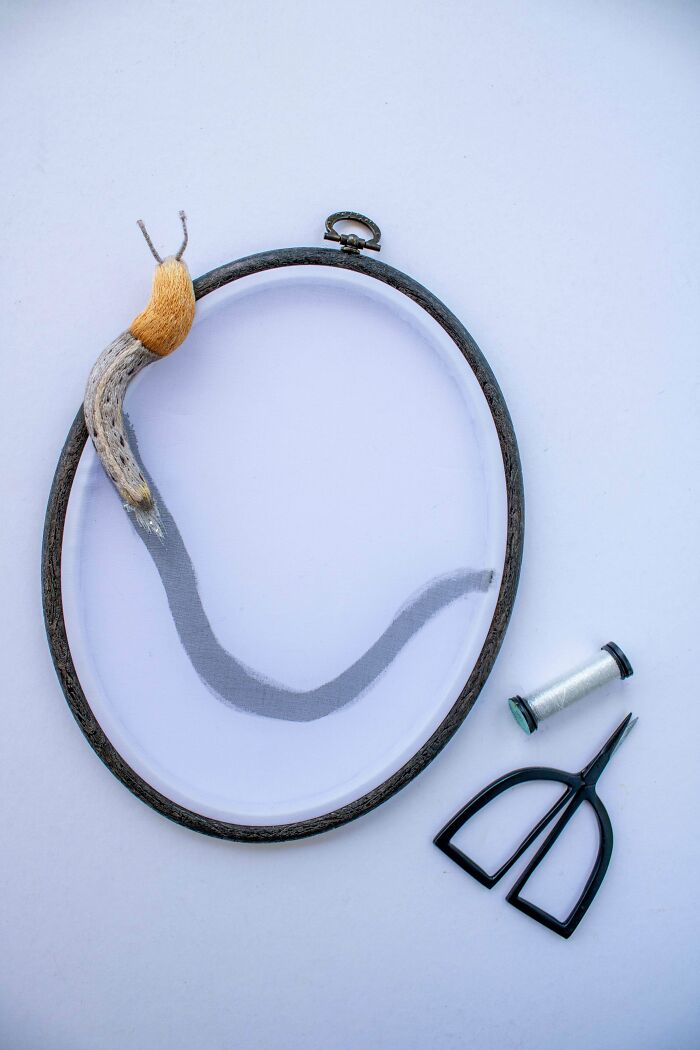 Embroidery hoop featuring a realistic snail created with threads, accompanied by scissors and a spool of white thread.