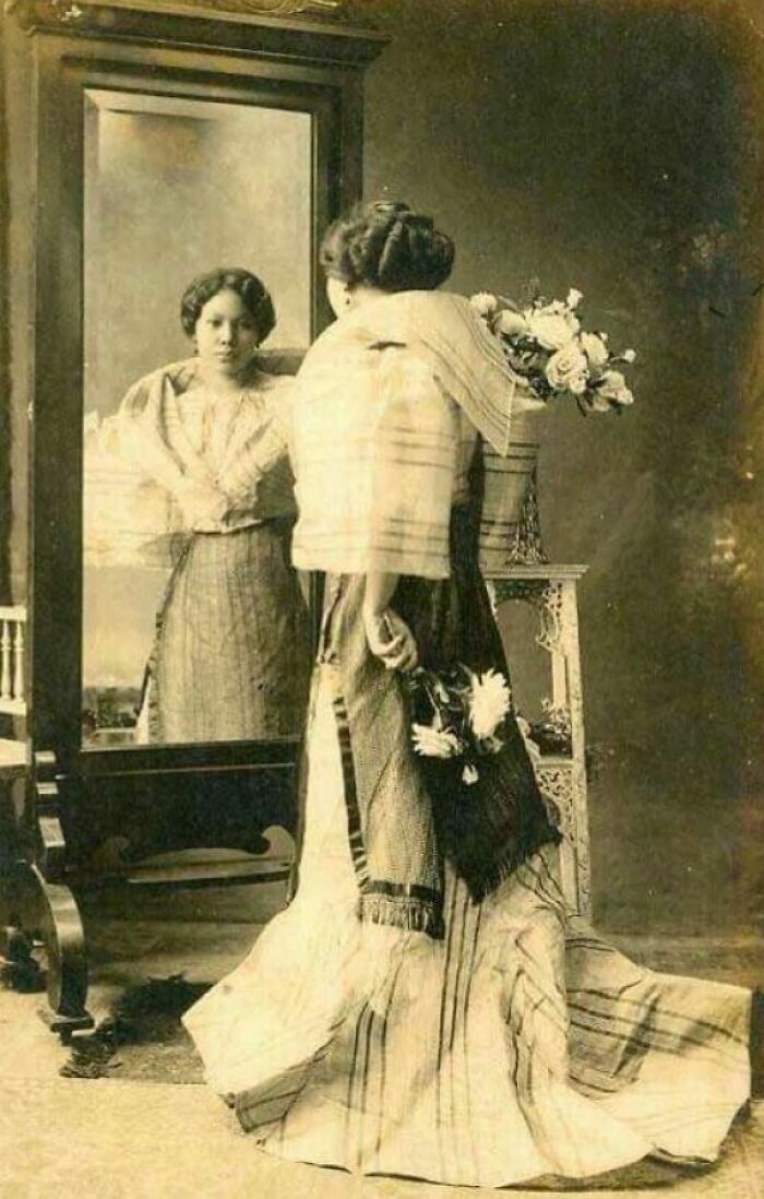 Filipina in traditional 1890s attire holding flowers, looking into a mirror in a vintage room in the Philippines.