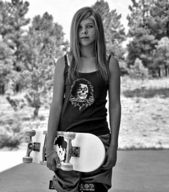 Teen girl holding a skateboard, wearing graphic tank top, standing outdoors reflecting on her regrettable past in old photo.