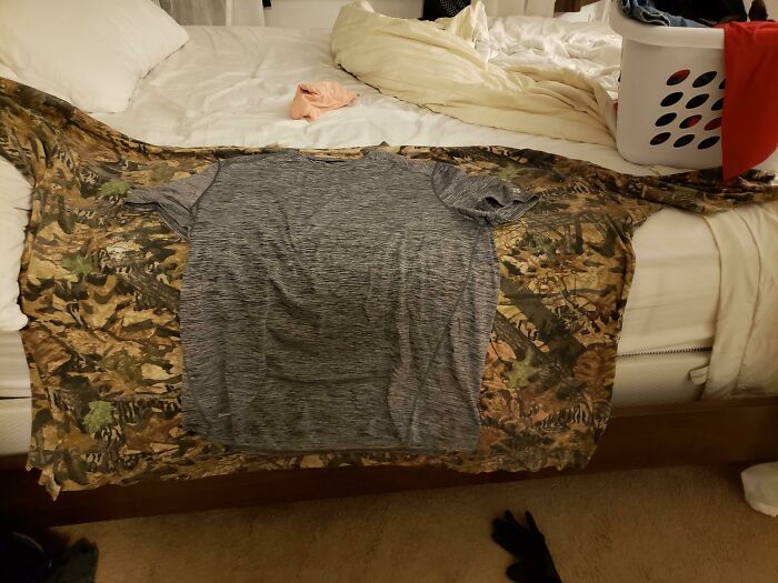 Gray t-shirt laid out on a camo jacket on a bed, demonstrating size comparisons for visual interest.