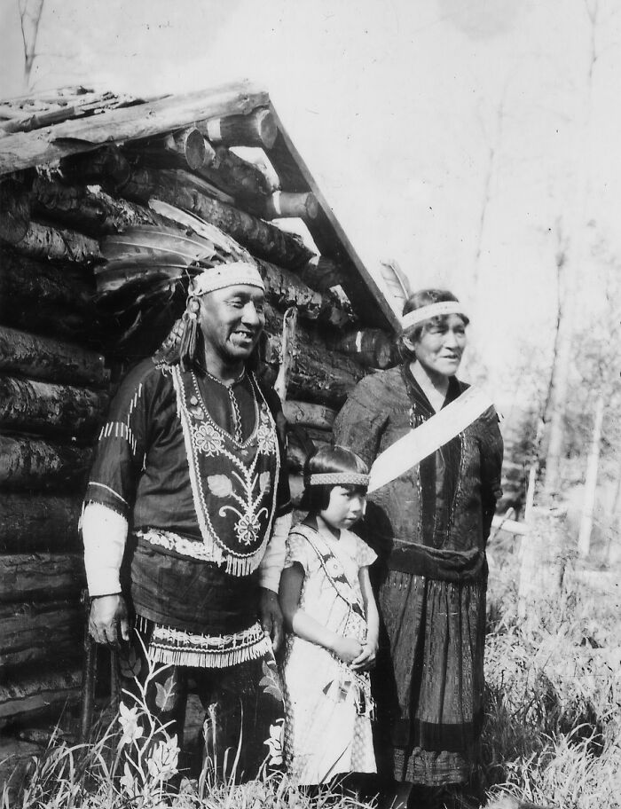 Vintage Native American family dressed in traditional clothing standing outdoors near a log cabin in a historical black and white photo.