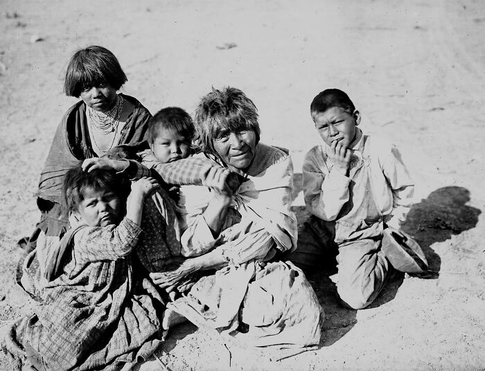 Vintage Native American family photo showing an elder woman and children sitting together outdoors in traditional attire.